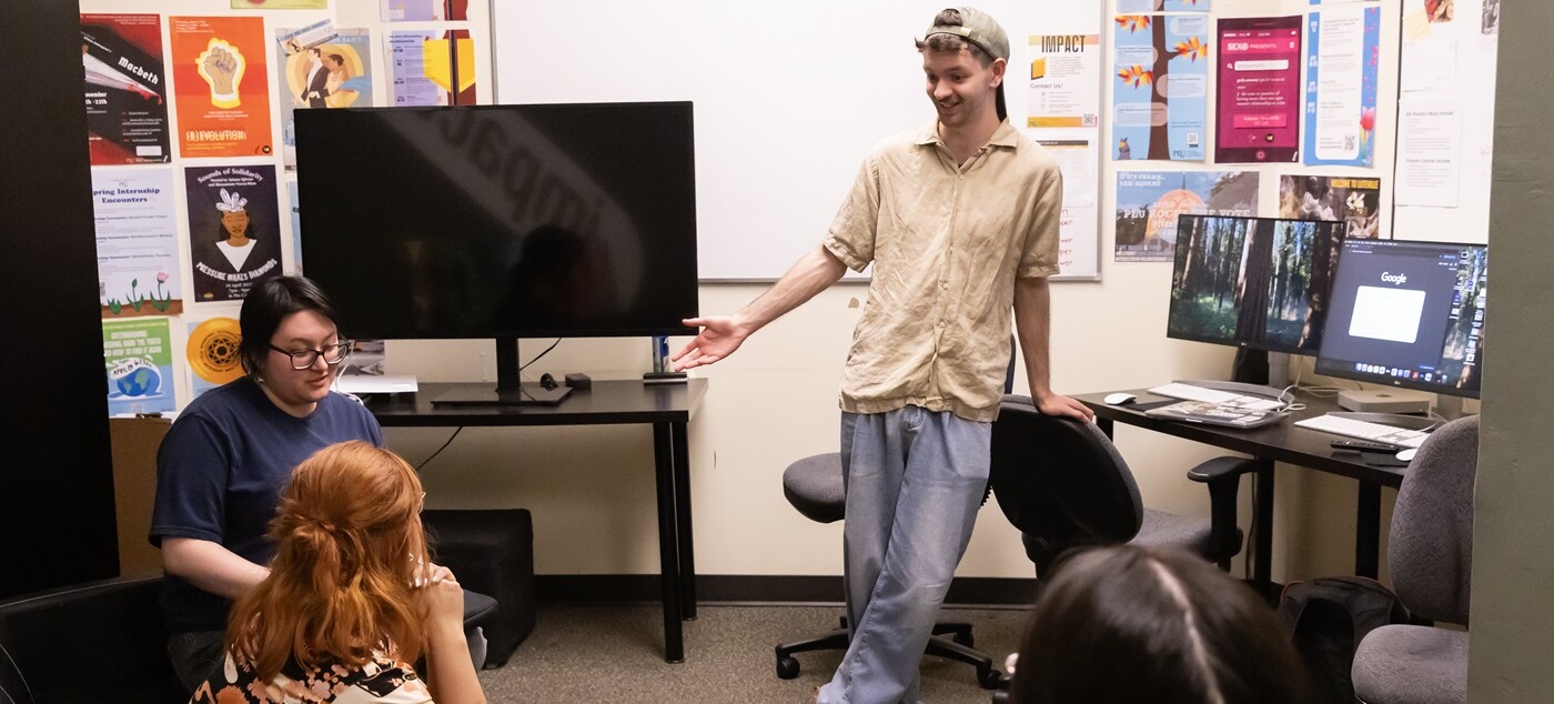 Ivan Wilson, stands leaning against the back of a chair in PLU's Impact Office. He's chatting with two other students who are sitting. In the room are computers, a TV monitor and multiple posters on the wall.