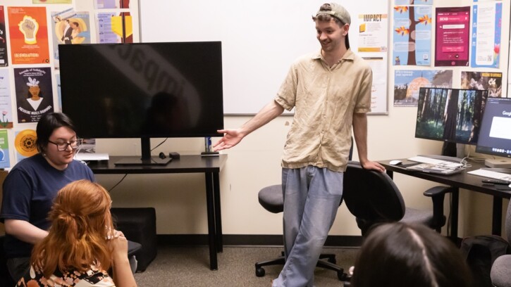 Ivan Wilson, stands leaning against the back of a chair in PLU's Impact Office. He's chatting with two other students who are sitting. In the room are computers, a TV monitor and multiple posters on the wall.