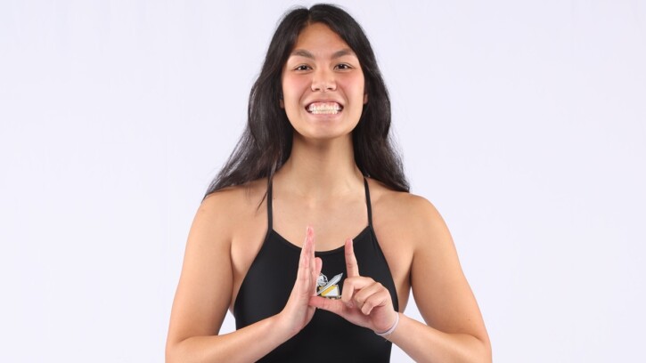 Student-athlete Matthea Gaines stands in front of a white background wearing a black PLU swimsuit. She is signing something in ASL.