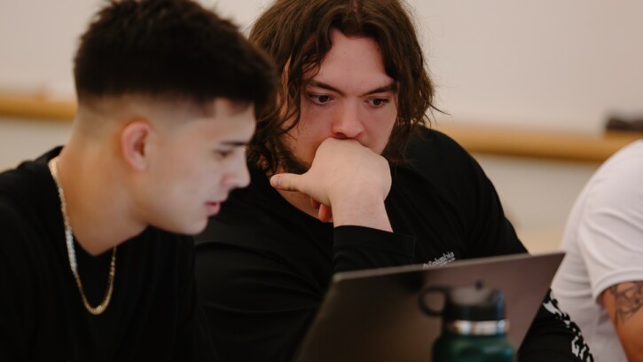 Two students sitting at a table next to each other look at a laptop together.