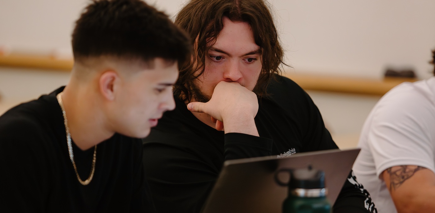 Two students sitting at a table next to each other look at a laptop together.