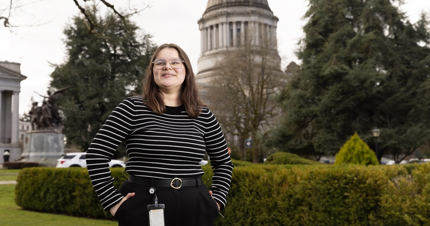 Cady Wood, dressed in a black and white striped shirt, and black pants with a badge attached to her belt, stands with her hands in pockets outside, looking just beyond the camera. Behind her are bushes, trees and the large white and grey Washington State capitol building.