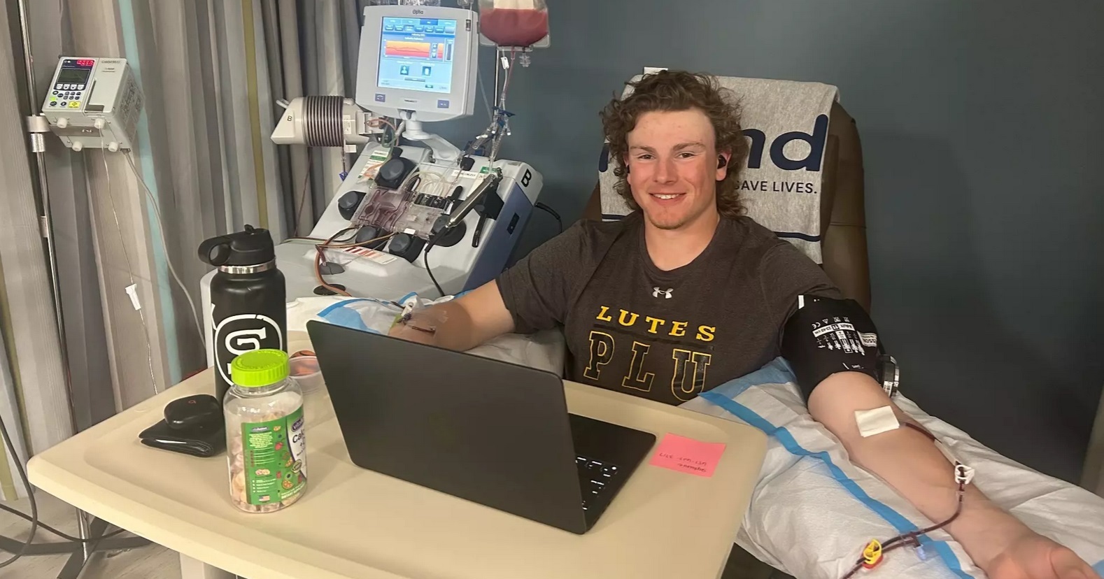 Carson Burke, in a PLU t-shirt, sits up in a hospital bed with his laptop open in front of him. His left arm is propped up on a pillow with an IV inserted, as he donates stem cells.