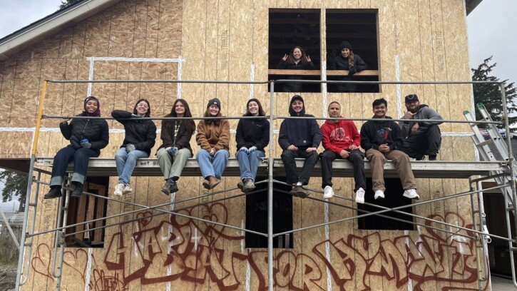 A group of students from the J-Term on the Hill Class sit on scaffolding against a house being built with Habitat for Humanity. Below is graffiti on the unpainted house that reads 'Habitat for Humanity'.