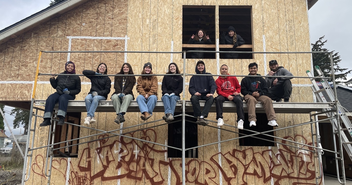 A group of students from the J-Term on the Hill Class sit on scaffolding against a house being built with Habitat for Humanity. Below is graffiti on the unpainted house that reads 'Habitat for Humanity'.