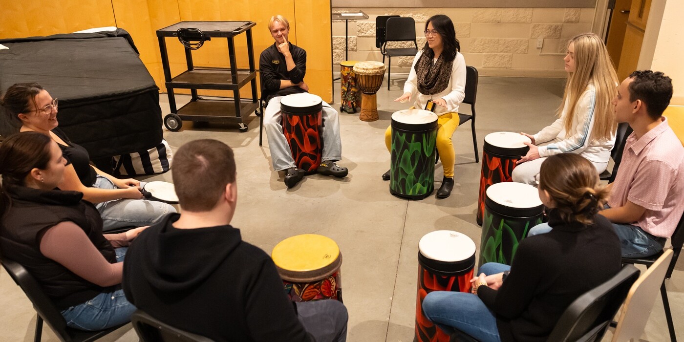 A psychology of music class, led by Assistant Professor of Music and Director of Music Therapy sit in a circle, each with a large brightly colored drum in front of them.