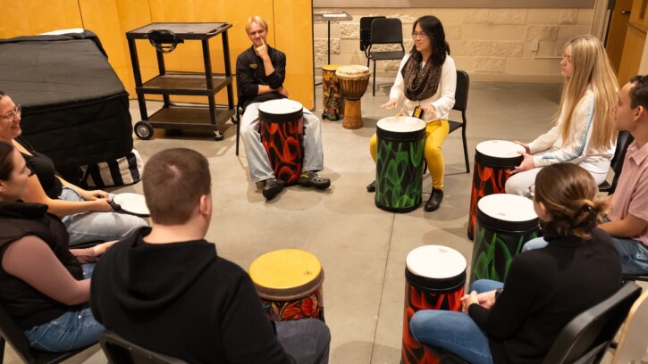 A psychology of music class, led by Assistant Professor of Music and Director of Music Therapy sit in a circle, each with a large brightly colored drum in front of them.