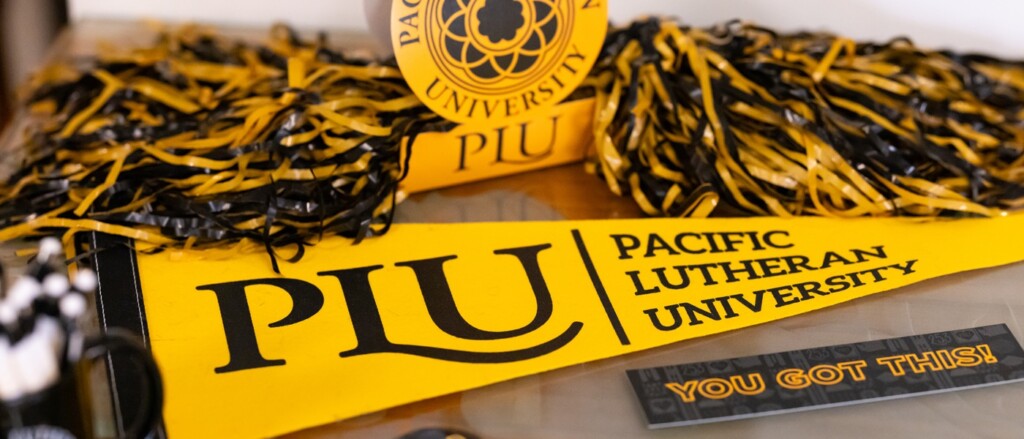 A yellow PLU pennant that reads PLU - Pacific Lutheran University, sits on a table, surrounded by other black and yellow PLU stickers and pom poms