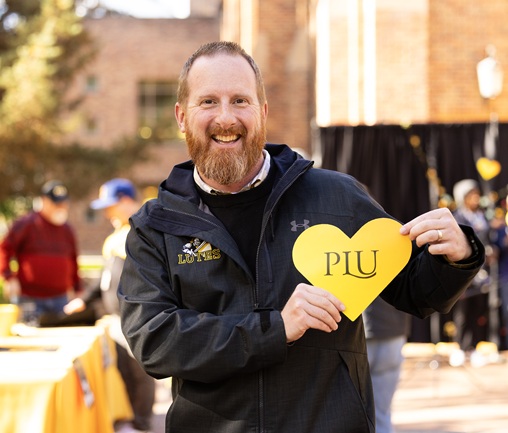 Director of Athletics & Recreation, Mike Snyder, wearing a Lutes jacket holds up a paper yellow heart that reads 'PLU' during Bjug Day on campus.