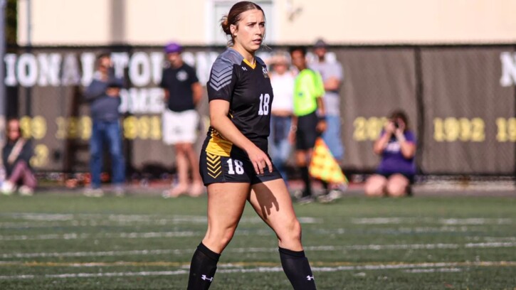 Carmen Kiewert, wearing black and yellow PLU women's soccer uniform walks the field on a sunny game day.