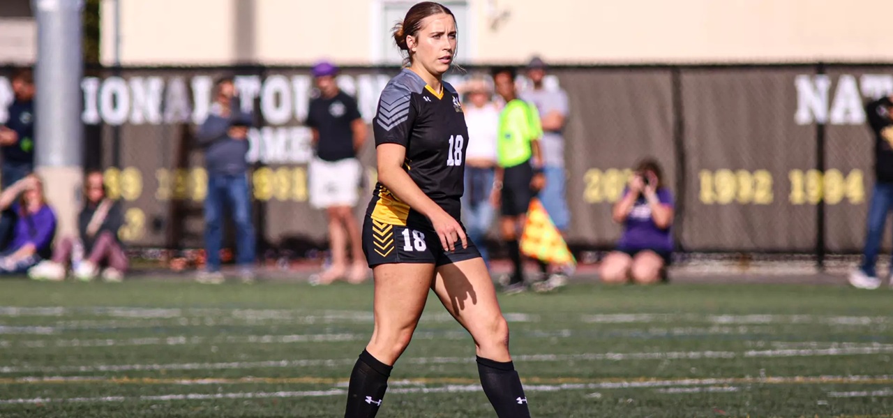 Carmen Kiewert, wearing black and yellow PLU women's soccer uniform walks the field on a sunny game day.