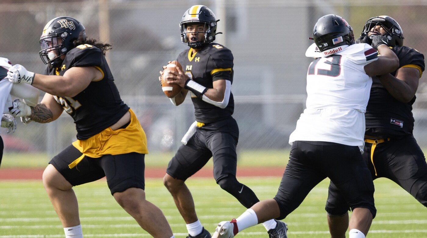 A PLU quarterback holds the ball, looking for a play while linemen on either side grapple with opponents during a game on a sunny day.