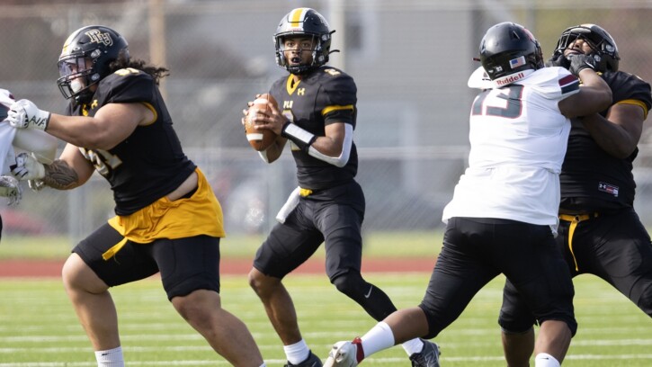 A PLU quarterback holds the ball, looking for a play while linemen on either side grapple with opponents during a game on a sunny day.