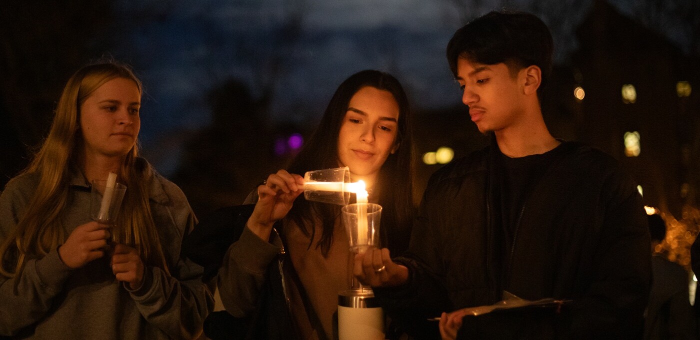 Three students stand side-by-side outside in the evening at PLU's annual Celebration of light. Each are holding a candle in a cup - the candle held by the student on the left is unlit, while the student in the middle is lighting their candle from the student on the right.