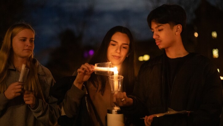 Three students stand side-by-side outside in the evening at PLU's annual Celebration of light. Each are holding a candle in a cup - the candle held by the student on the left is unlit, while the student in the middle is lighting their candle from the student on the right.