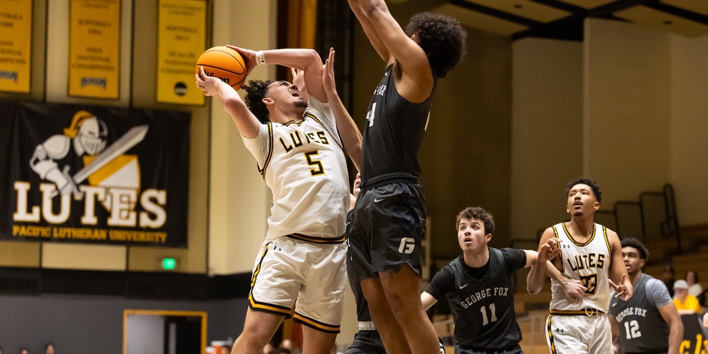 A PLU Men's Basketball player jumps toward the hoop in the PLU Olson Gym. A player on the opposing team jumps to try to block. In the background are more players and and Lutes sports banner hanging on the wall.