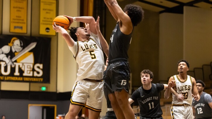 A PLU Men's Basketball player jumps toward the hoop in the PLU Olson Gym. A player on the opposing team jumps to try to block. In the background are more players and and Lutes sports banner hanging on the wall.
