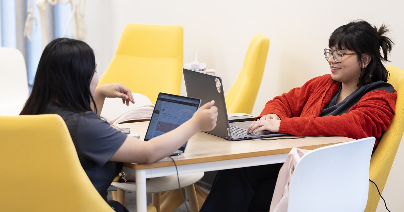 Two students sit at a table in the PLU library. Both are in bright yellow chairs and have laptops open in front of them.