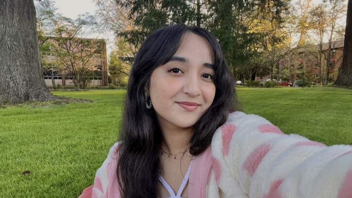 PLU student Mahnoor Iftikhar takes a selfie. In the background is a grassy field, brick buildings and trees on the PLU campus.