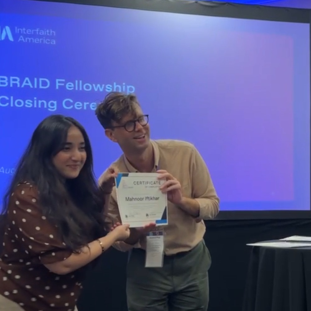 Mahnoor and another person stand next to each other holding a certificate. In the background is a projector screen that reads 'Interfaith America - BRAID Fellowship Closing Ceremony'