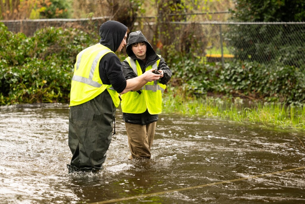 Two students in rain gear, waders and yellow reflective vests stand mid-creek to measure flow rates in Clover Creek during an Environmental Methods class.