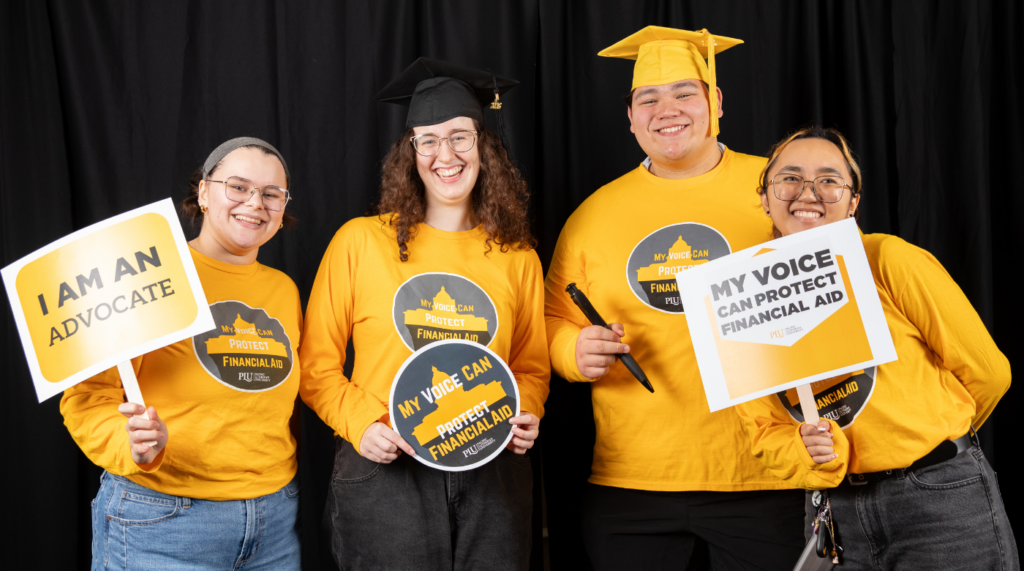 Four students hold signs and look into the camera and smile