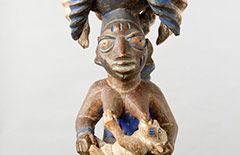 This wooden sculpture depicts a kneeling mother with a nursing child in her arms, while carrying another child on her back. This maternal depiction reflects the revered role of Yoruba mothers as creators of new life. But her kneeling posture is also an expression of respect and submission and reflects the revered status of the deities. The carved bowl on top of her head is utilized in divination rituals, where guidance is asked for from Ifa, the God of divination.