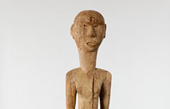 This is a Bongo grave post with a male figure, which is carved out of a single piece of wood. Details include small metal pieces, which are found on the head and chest and represent tattooing that occurs in this society. Additionally, there is also a piercing on the stomach near the bellybutton.
