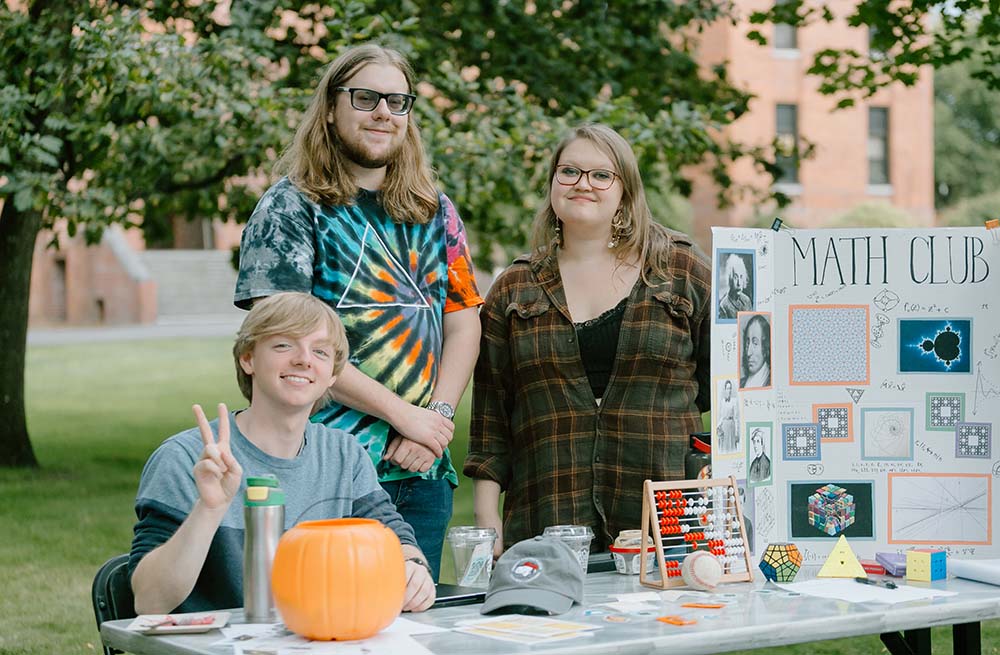 Three students sit and stand next to a poster about Math Club at the fall 2023 PLU involvement fair.