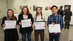 Students receive awards for their artwork during a gallery show. Four students standing and smiling at camera hold up their certificates.