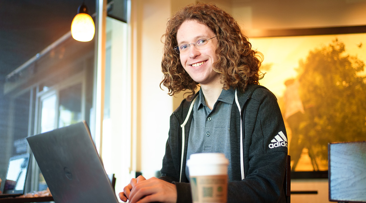 PLU alum Matthew Conover sits smiling at his laptop computer in a coffee shop with a cup of coffee a few inches away.