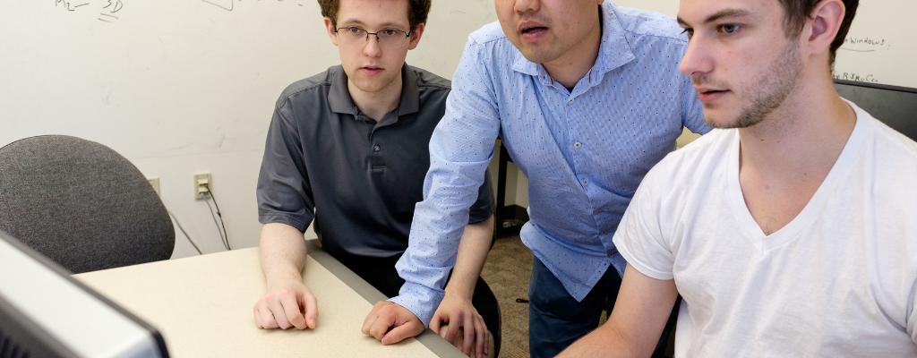 two students (seated) and a professor (standing) look at a shared computer screen.