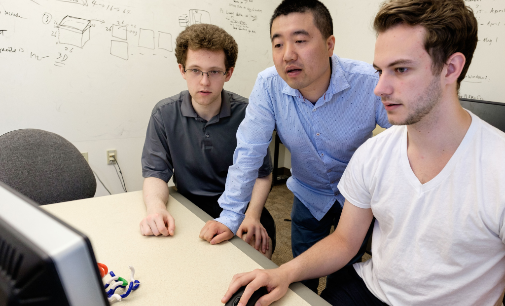 two students (seated) and a professor (standing) look at a shared computer screen.