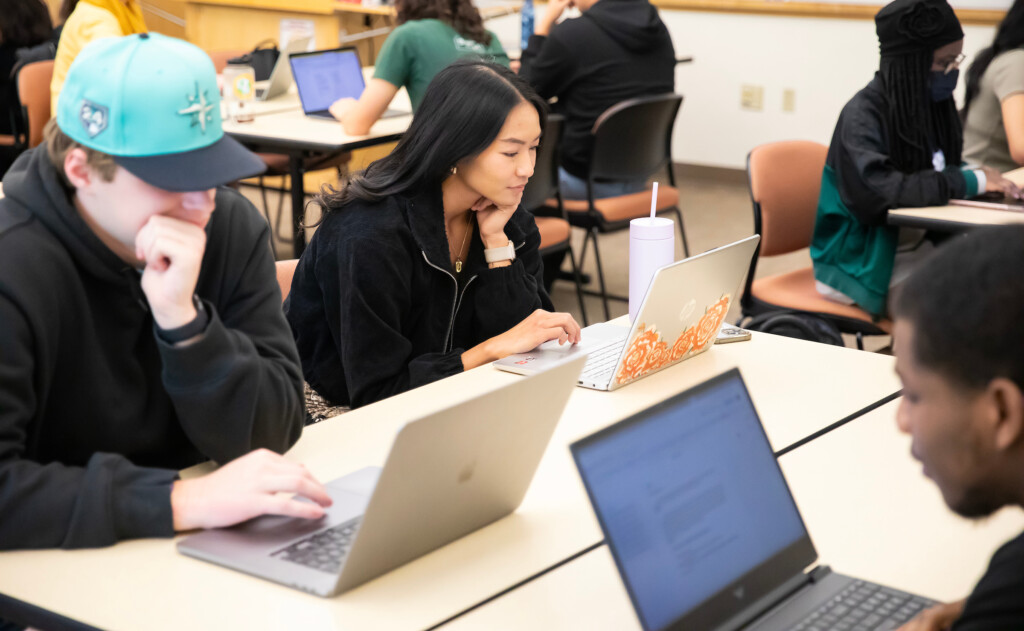 Three students work on their laptops in a classroom setting.