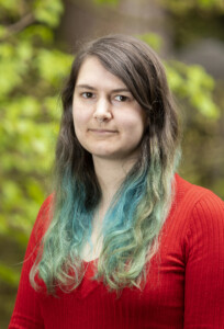 Headshot of a white person with long brown and teal hair in a red sweater, with trees in the background.