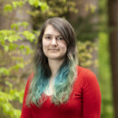 Headshot of a white person with long brown and teal hair in a red sweater, with trees in the background.