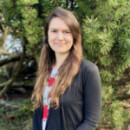 White person with long brown hair in a black cardigan standing in front of trees.