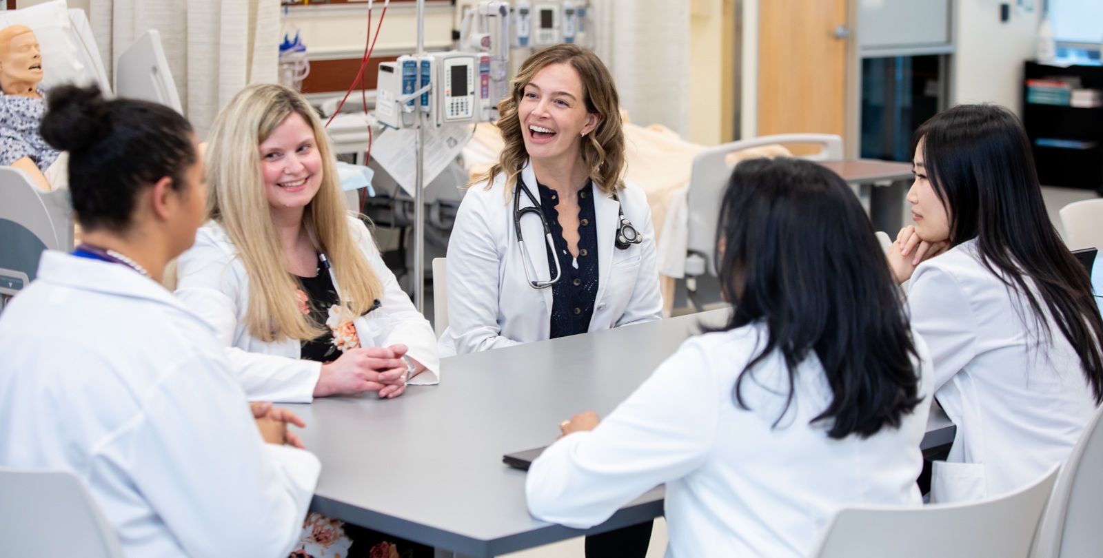 Web Banner - DNP 01 A group of DNP students in white lab coats sit around a table chatting in the PLU Nursing Simulation Center