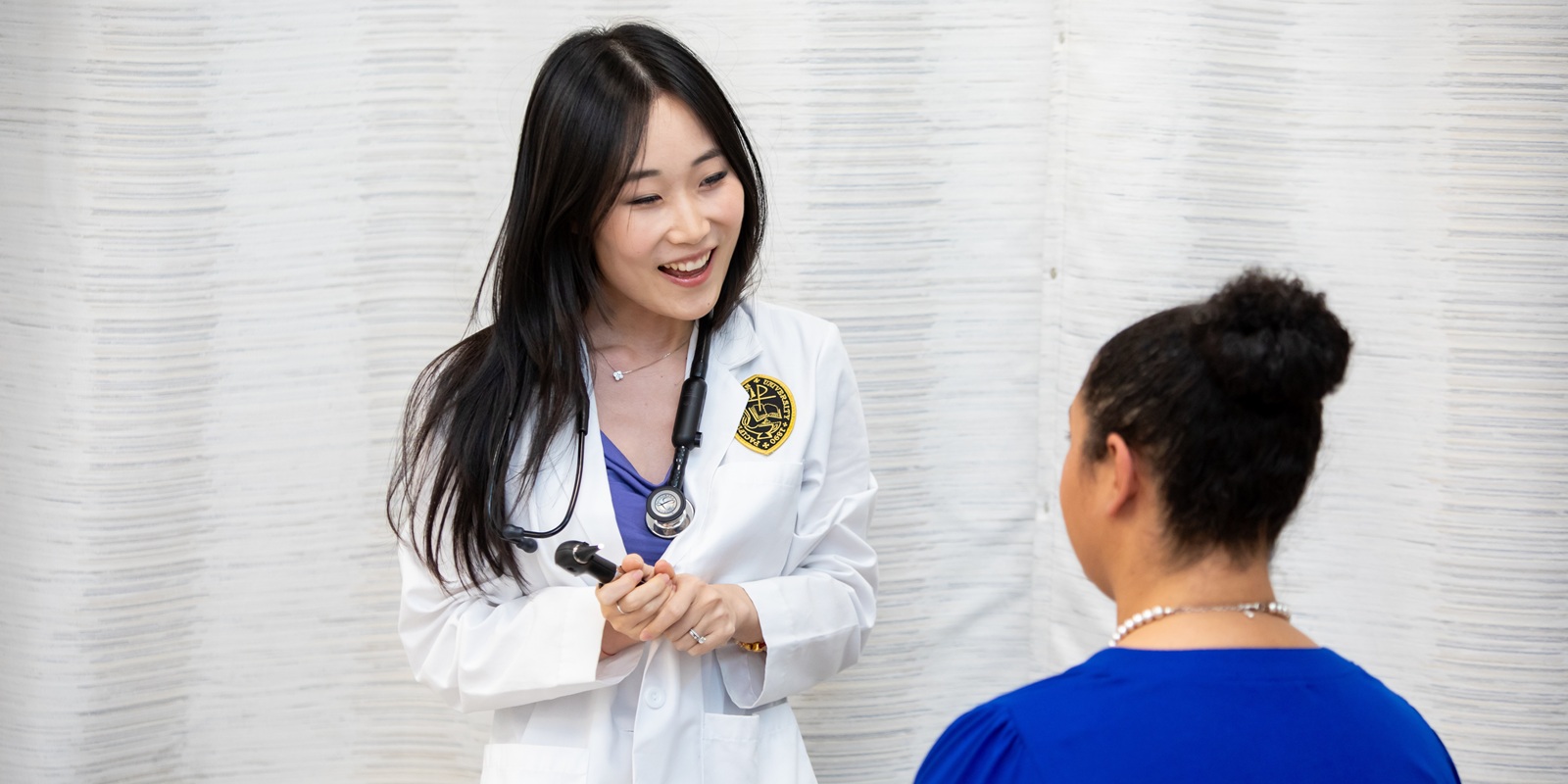 Web Banner - DNP 02 A PLU DNP student in a white lab coat and wearing a stethoscope around her neck stands to chat with a sitting patient who has their back to the camera.