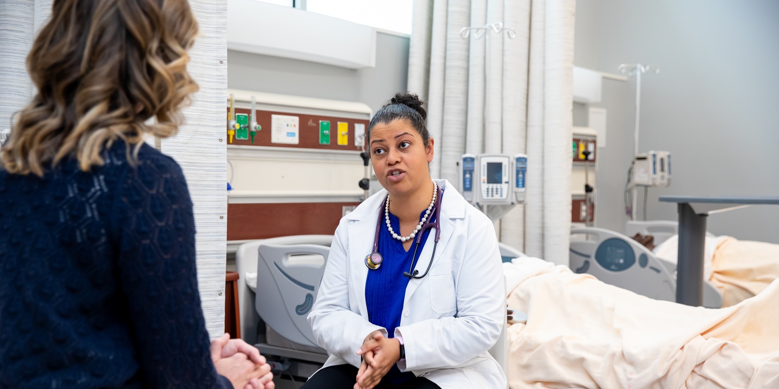 Web Banner - DNP 03 A PLU DNP student wearing a white lab coat and stethoscope around her neck, sits in PLU's Nursing Simulation Center with a patient bed behind her. In front of her is a patient sitting, with their back to the camera.