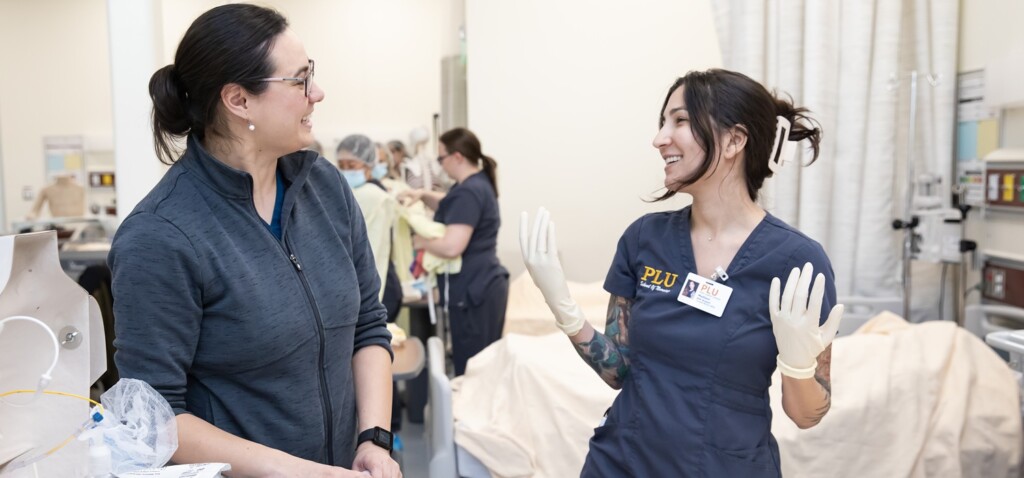 Web Banner - Nursing 03 A PLU nursing instructor and student wearing latex gloves and blue-grey PLU School of Nursing scrubs smile at each other while in the PLU clinical simulation lab.