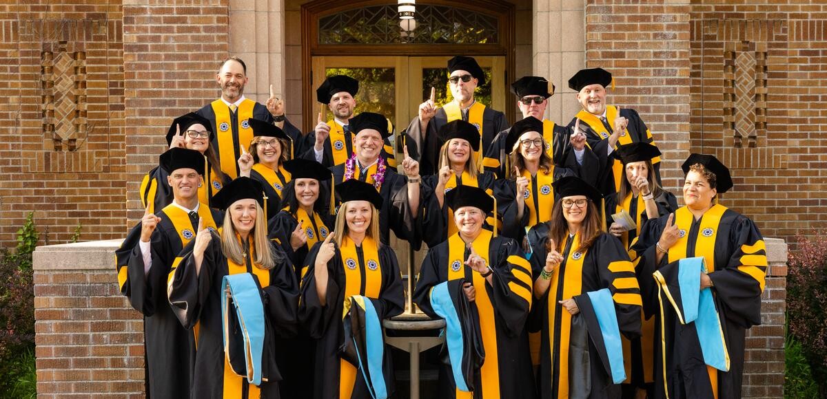 20260521_SB_Professional-Studies-Commencement_015-medium Group of Ed.D graduates in black robes and yellow sashes posing before brick building.