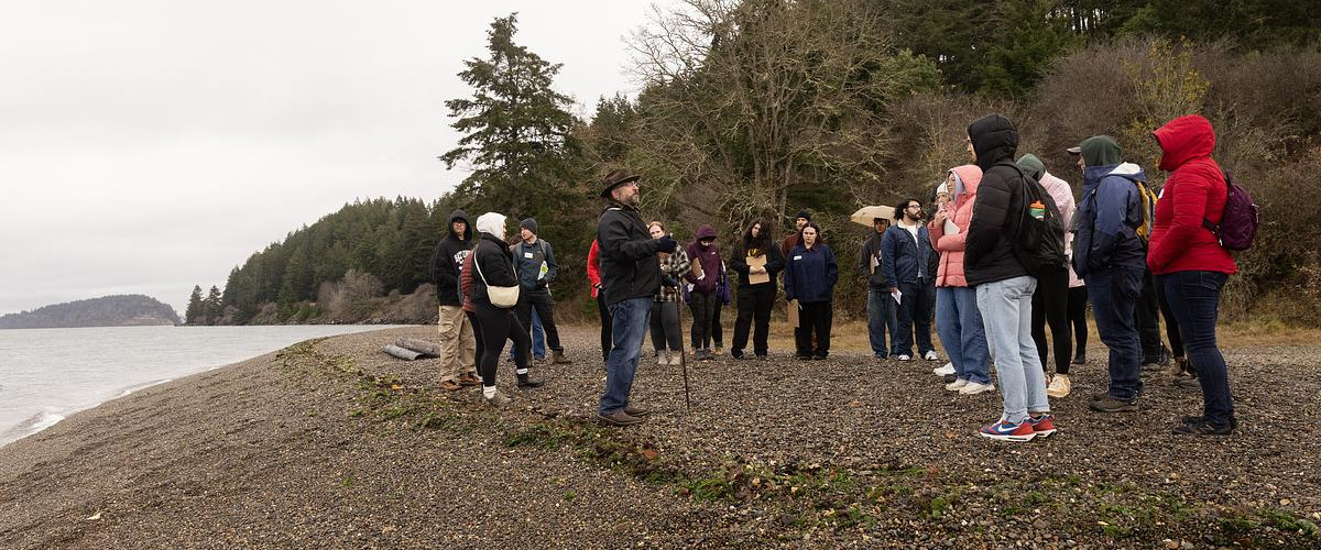 Students standing on a rocky beach with trees behind them. They are looking out at a guide who is standing near the water and speaking to them.