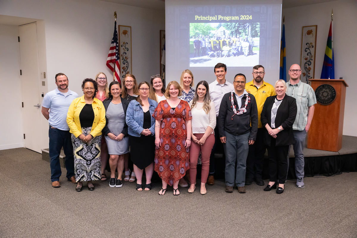 Several principals standing in front of a screen that says "Principal Program 2024." Several flags are also behind them, along with a podium that bears Pacific Lutheran University's logo.