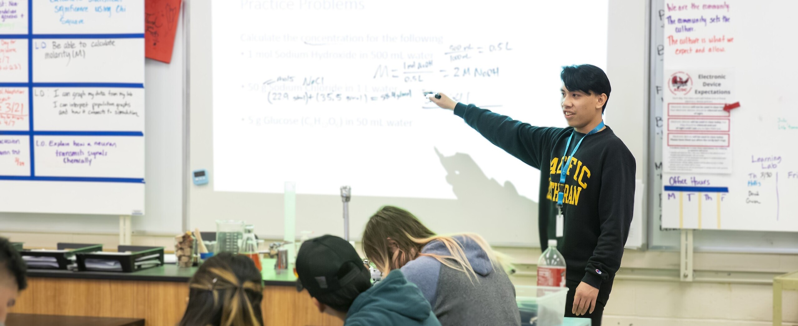 student teaching class header teacher standing in front of class giving a lecture. They are writing on a white board and the students have their heads down, presumably taking notes.
