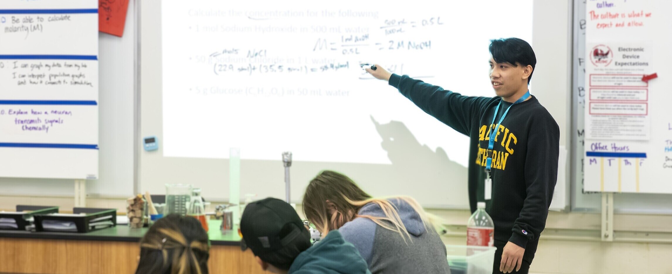 teacher standing in front of class giving a lecture. They are writing on a white board and the students have their heads down, presumably taking notes.