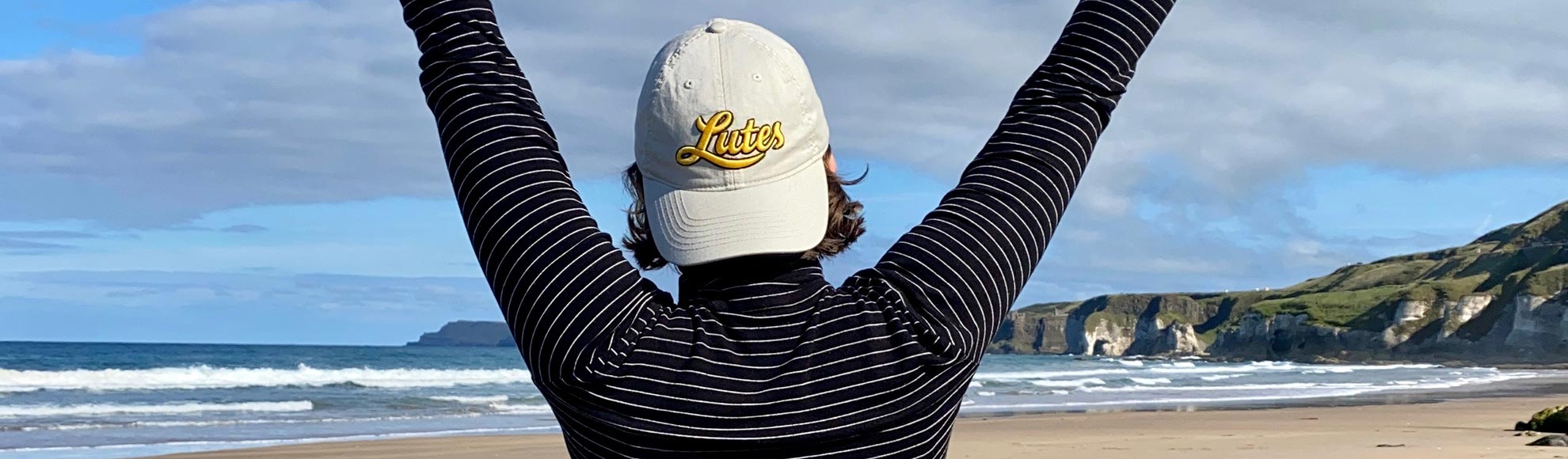 A PLU student in the foreground, wearing a PLU hat, with the ocean and a beach in front of them.