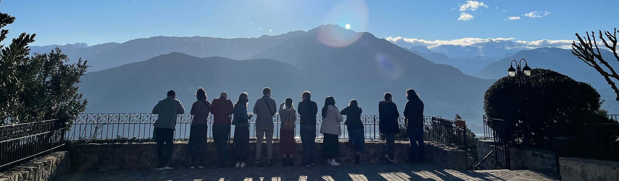 Several people standing near a railing with their backs to the camera. There is a beautiful mountain range in the background, with clouds floating behind them.
