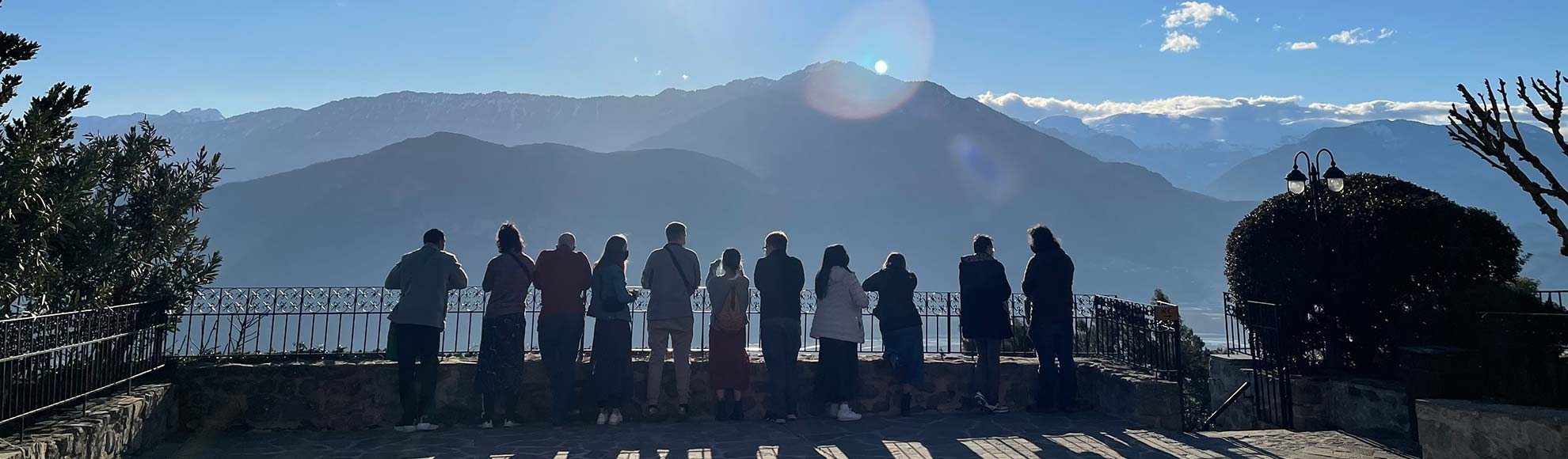 Several people standing near a railing with their backs to the camera. There is a beautiful mountain range in the background, with clouds floating behind them. Several people standing near a railing with their backs to the camera. There is a beautiful mountain range in the background, with clouds floating behind them.