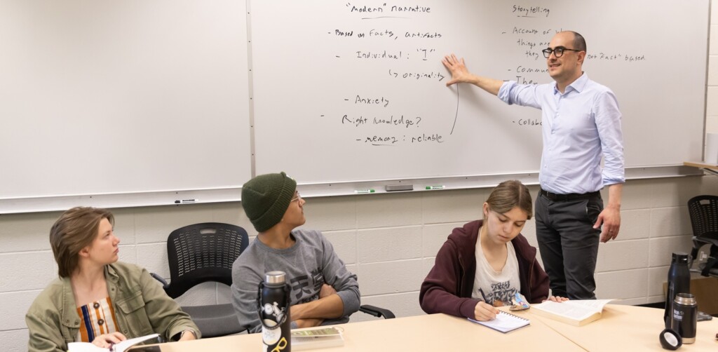 Resident Assistant Professor Christian Gerzso leads a roundtable discussion in his IHON 112 course titled “Liberty, Power, and Imagination". The professor is standing next to and touching a white board with notes on it, while in front of him, three students sit at a table with books, notebooks and water bottles in front of them.